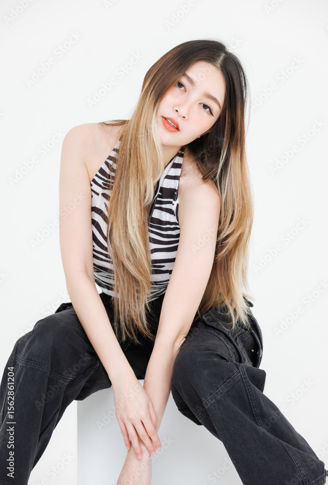 Photo trendy fashion asian attractive pretty young woman model poses confidence wear zebra print halter top, black cargo pants streetwear style sitting on cube box look at camera in white background. Photo trendy fashion asian attractive pretty young woman model poses confidence wear zebra print halter top, black cargo pants streetwear style sitting on cube box look at camera in white background.