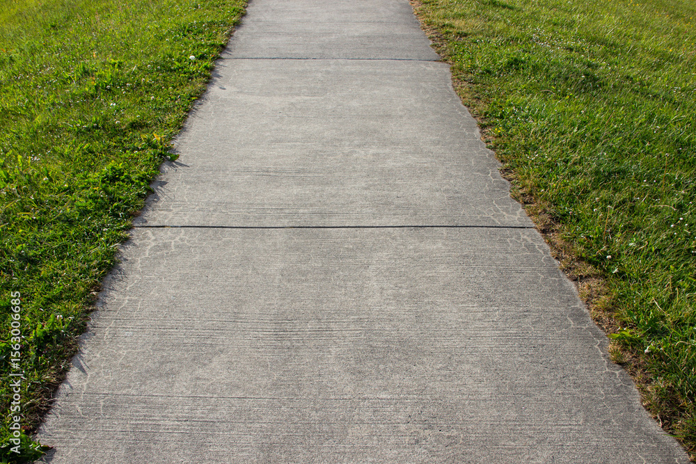 Concrete walkway bordered by green lawn on both sides on a sunny day. Clean modern garden path, urban or suburban landscaping. Concrete walkway bordered by green lawn on both sides on a sunny day. Clean modern garden path, urban or suburban landscaping.