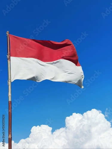 The red and white Indonesian flag on a bamboo pole flutters in the wind against a backdrop of cloudy blue sky.