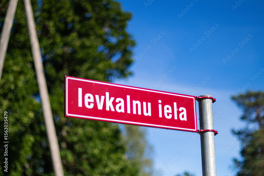 Fototapeta premium Close-up of a red Latvian street sign labeled “Ievkalnu iela,” captured outdoors against a clear blue sky and greenery.