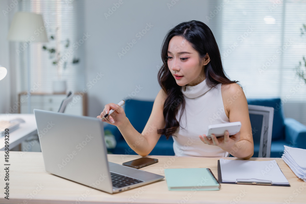 Obraz premium Young professional woman multitasking at her desk, using a laptop and calculator for business tasks. Modern office setting with natural light and organized workspace