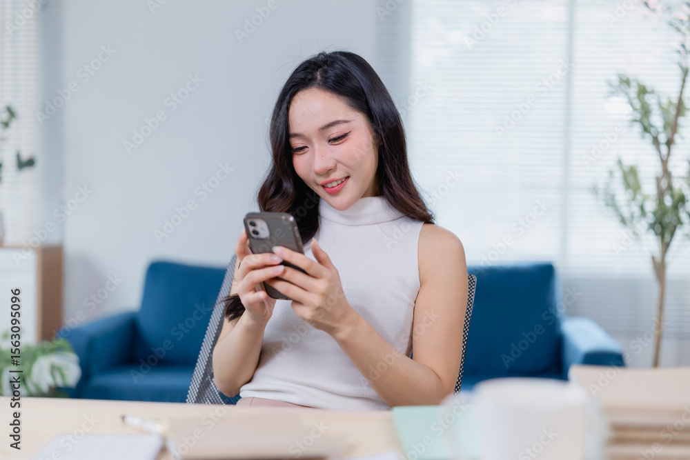 Young woman sits at a desk in a modern office, smiling as she engages with her smartphone. The bright, airy space includes a blue sofa and natural light from large windows Young woman sits at a desk in a modern office, smiling as she engages with her smartphone. The bright, airy space includes a blue sofa and natural light from large windows