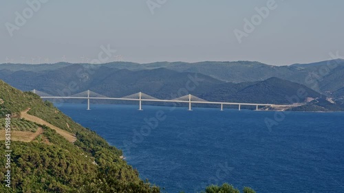 Panoramic view of Peljesac Bridge connecting Croatian mainland and peninsula across the deep blue Adriatic sea with forest hills wind turbines and clear sky modern European infrastructure