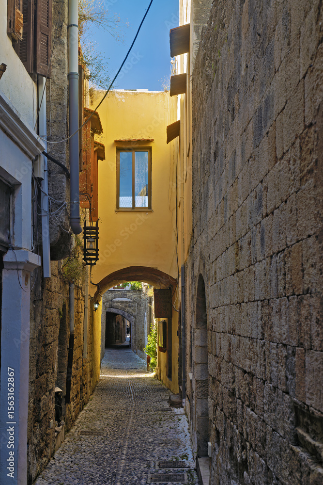 Fototapeta premium narrow alleyway paved with stone tiles and flanked by aged stone and stucco buildings stone arches span across the alley overhead in old town of Rhodes, Greece