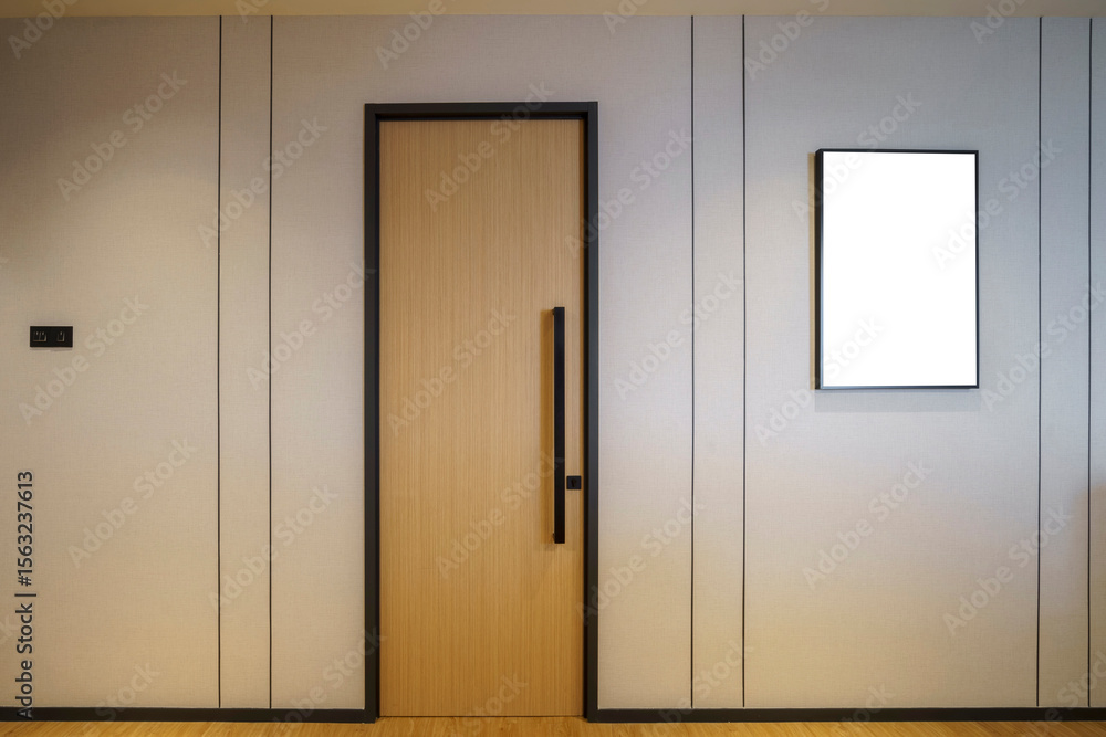 Fototapeta premium A modern light wooden door with a black frame and a long vertical handle, set against a gray upholstered wall with an empty frame and a socket.