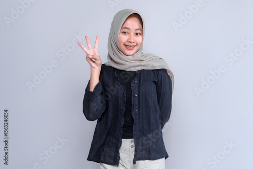Portrait of a cheerful young Asian woman wearing a light grey hijab and black blouse, smiling confidently while showing three fingers gesture against a plain background.