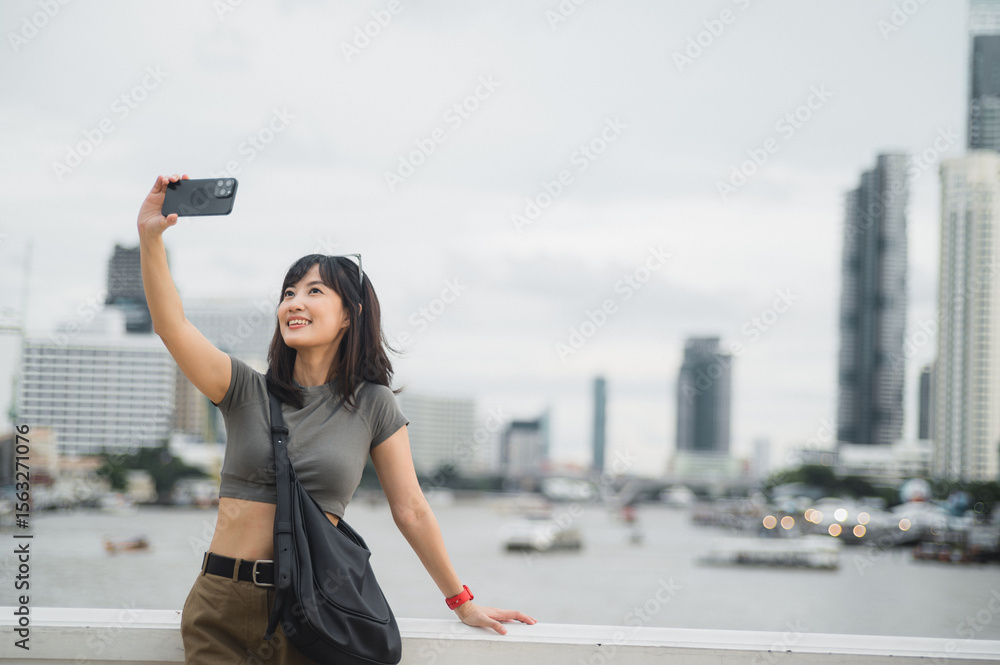 Fototapeta premium Beautiful young southeast asian woman blogger and world travel explorer in 30s taking happy selfie with smartphone at Chao Phraya River in Bangkok. summer tourism concept