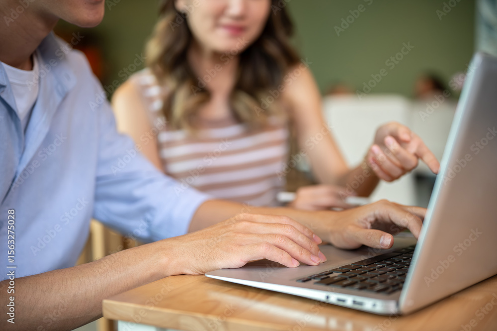 Close up of asian man typing on laptop with woman pointing sits at wooden table in cafe or canteen. Close up of asian man typing on laptop with woman pointing sits at wooden table in cafe or canteen.