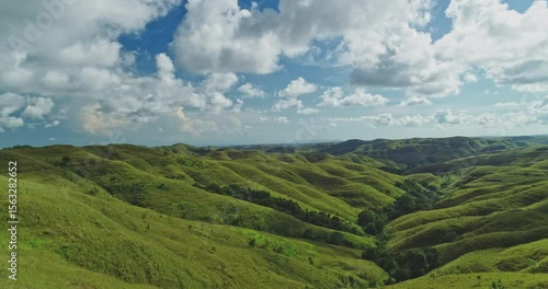 Young woman standing on a cliff with arms outstretched, embracing the breathtaking landscape of rolling green hills and blue skies on Sumba Island, Indonesia