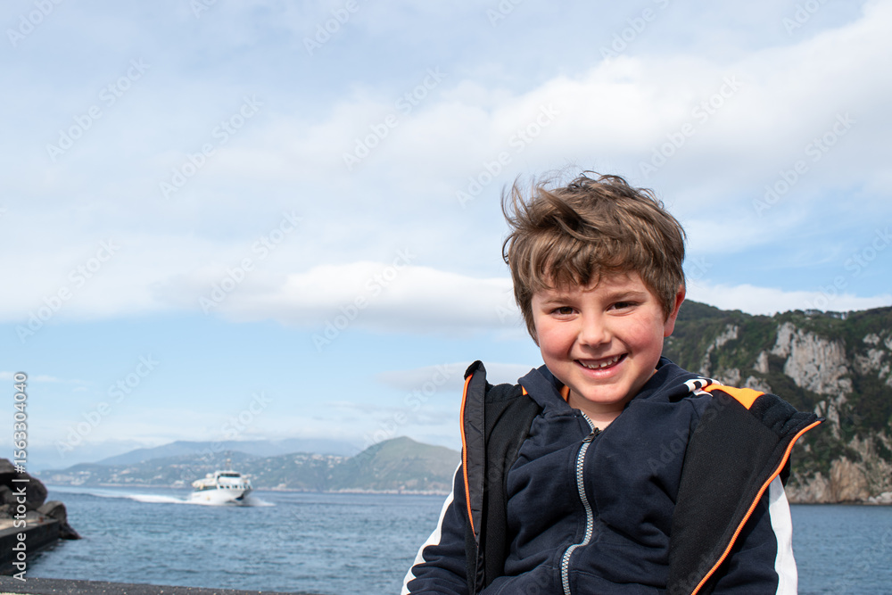 Fototapeta premium Smiling happy boy looking at camera with sea bay and boat in background.Text space.