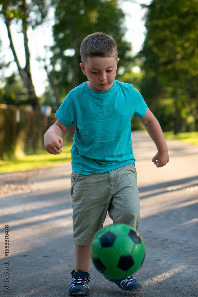 Obraz premium A 5-year-old boy is joyfully and passionately playing with a soccer ball on a quiet rural road