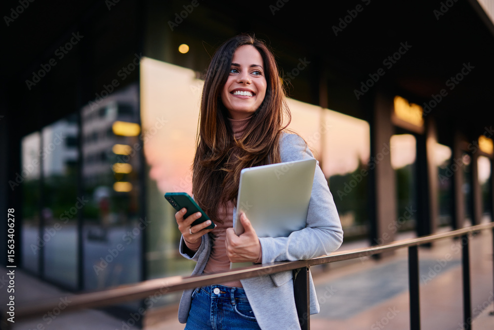 Obraz premium Smiling Professional Woman Holding Laptop and Phone in Modern Urban Setting