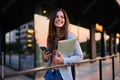 Bild auf Leinwand Smiling Professional Woman Holding Laptop and Phone in Modern Urban Setting