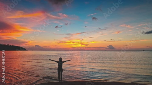 Silhouette of a young woman standing on a beach with open arms, embracing the vibrant and colorful sunset over the sea in Thailand, captured through stunning aerial drone footage