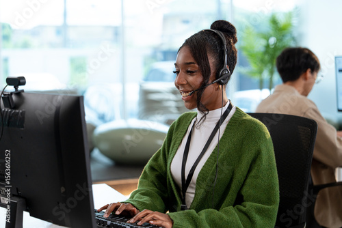 Photos A cheerful customer support representative wearing headset and green sweater is working at computer in modern office