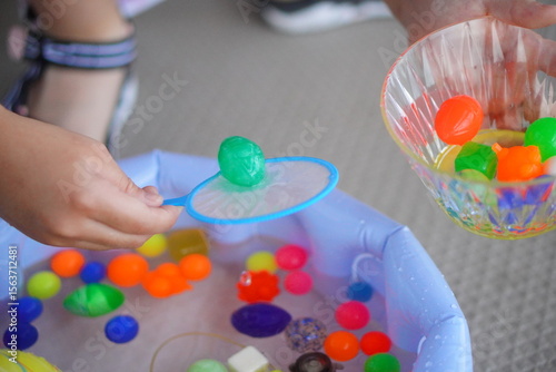 子供が屋外の水遊び用プールで鮮やかな緑色のスーパーボールを網に乗せ、横にある器に移そうとしている瞬間