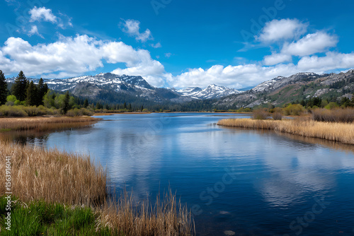 Serene blue lake reflecting white clouds against snow capped mountains and dry reeds image