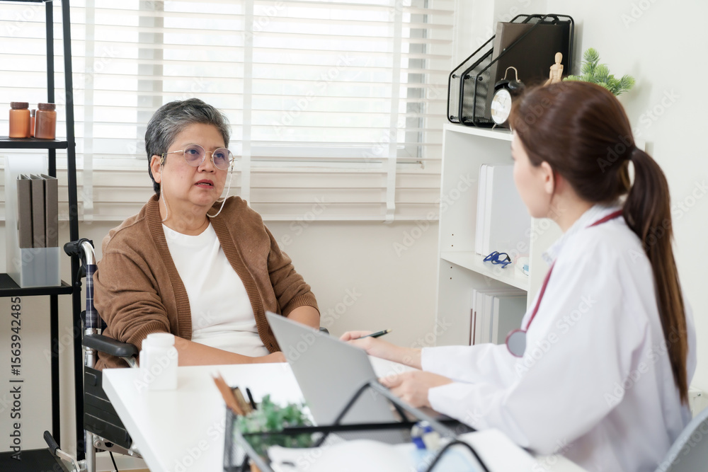 Fototapeta premium Elderly asian female sitting in wheelchair discusses symptoms of bone and joint pain with orthopedic doctor inside clinic office, having diagnostic consultation with notes and medicine bottle
