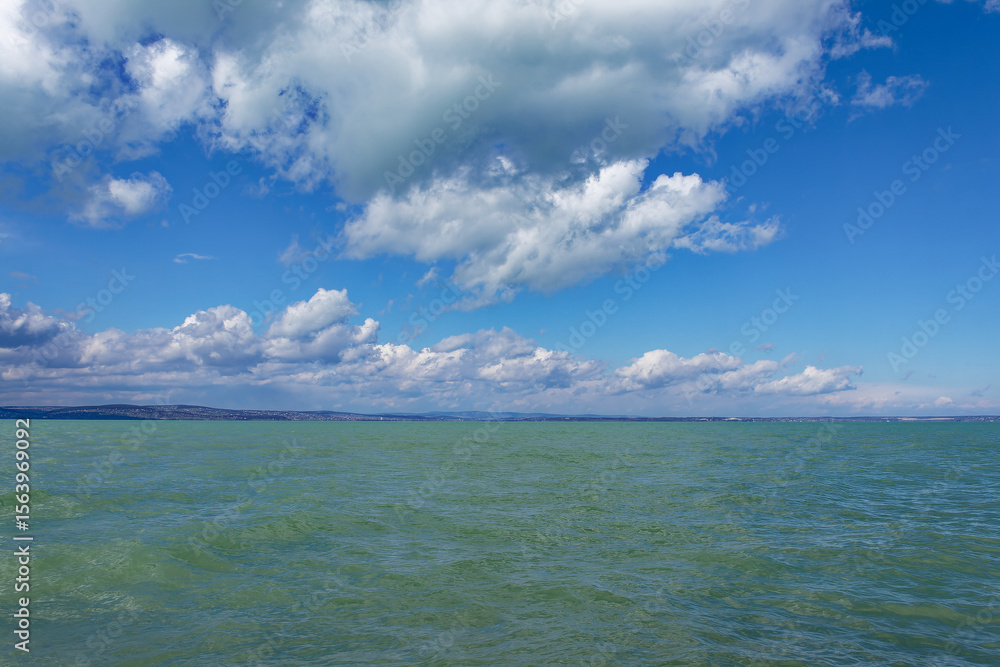 Obraz premium The lake stretches out beneath a clear blue sky, dotted with fluffy clouds on a bright day.