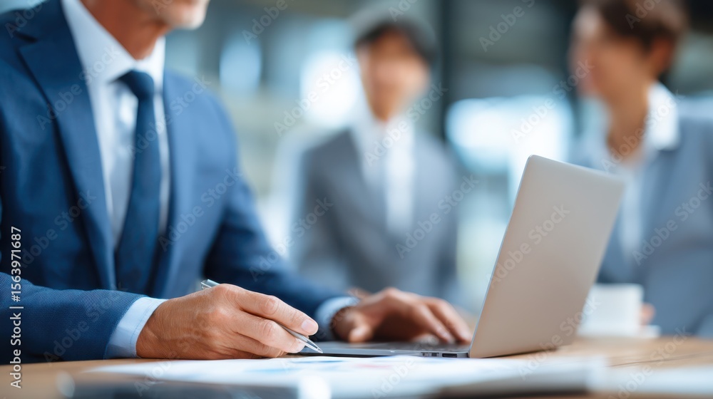 A professional individual works on a laptop while participating in a meeting with colleagues. The modern office environment enhances collaboration and discussion about project strategies A professional individual works on a laptop while participating in a meeting with colleagues. The modern office environment enhances collaboration and discussion about project strategies