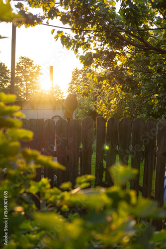 A clay flowerpot placed on top of a wooden fence, surrounded by glowing leaves and golden sunlight. Captures the cozy charm of rural life and summer evenings.