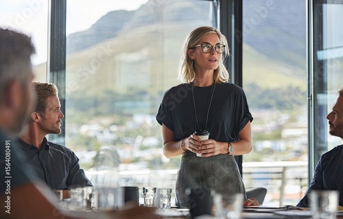 A woman in glasses presents to a small group seated around a table in a modern office with a scenic view.  She holds a coffee cup