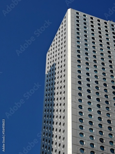 A striking tall building features a facade adorned with numerous circular windows, reflecting sunlight under a clear blue sky. The structure stands prominently in an urban setting.