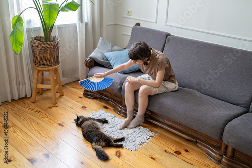 Schilderij op canvas Tired caring woman fan overheating cat on floor in hot apartment with no AC