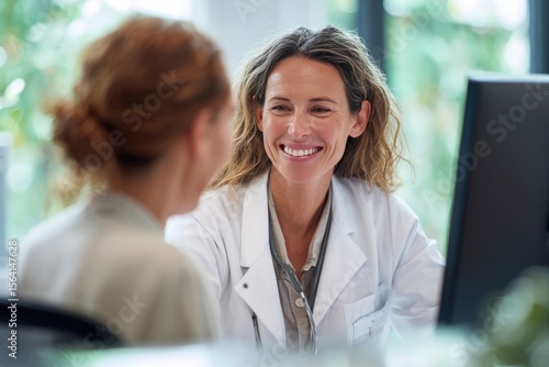 A smiling female doctor in a white coat sits at a desk, attentively listening to a female patient during a consultation in a bright, modern office