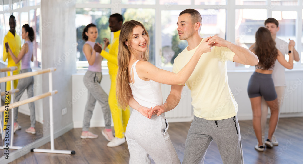 Fototapeta premium Positive young adult couple, attractive long-haired woman and sporty man dancing playful Latin dance bachata during group class in choreography studio. Active lifestyle concept