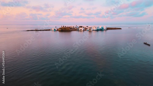 A group of small houses and a beach resort on top of a small island. The island is surrounded by blue waters with clouds in the sky above it. Location: Tunisia, Medenine , Zarzis.
