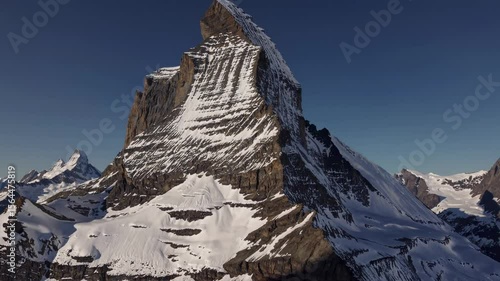 Aerial video captures a majestic snow-capped mountain peak under a clear blue sky, showcasing its rugged texture and towering presence from a high angle.