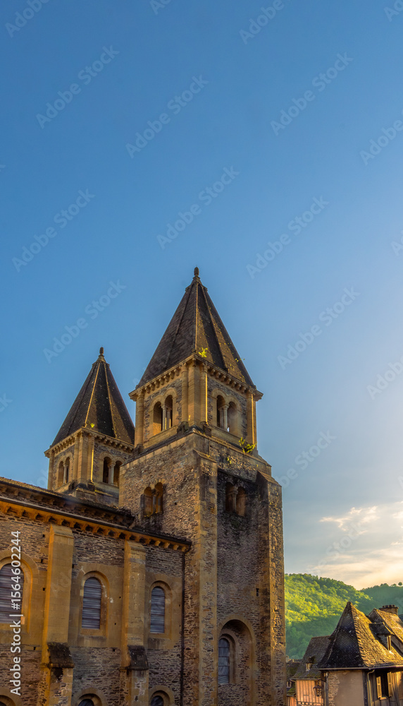 Obraz premium The twin spires of the ancient towers of Sainte-Foy Abbey and medieval houses are illuminated by the golden light of sunset under a clear blue sky in Conques, France.
