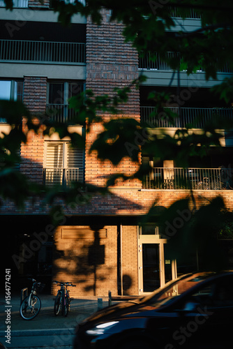 Vertical screen: sunlight illuminating brick facade of modern residential building at sunset with bicycles parked on sidewalk and car passing by, seen through foliage. Brick apartment exterior