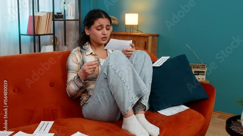Indian young woman sitting on sofa looking at bills, receipts, praying with eyes upward, sincerely asking for help. Arabian girl experiencing hope desperation, emotionally praying hoping for a miracle