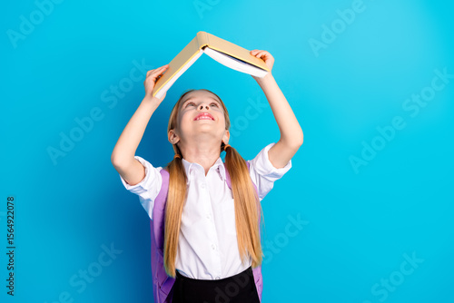 Bild auf Leinwand Happy schoolgirl holding a book on her head against a blue background, expressin