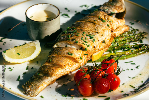 Grilled whole fish with asparagus, lemon wedge, cherry tomatoes, and sauce on plate