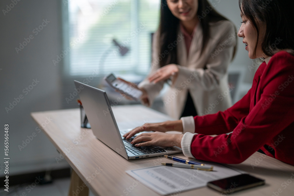 Two women are sitting at a desk with a laptop open Two women are sitting at a desk with a laptop open