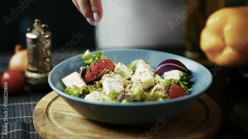 Hand seasoning fresh Greek salad in blue bowl