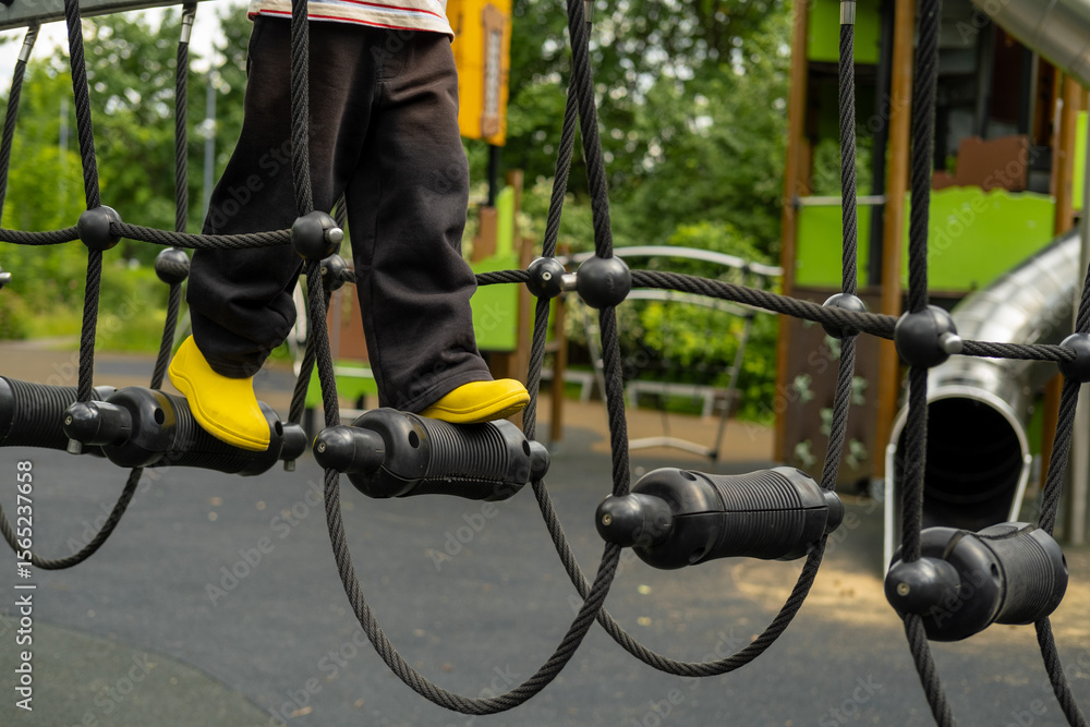 Obraz premium A child boy is stepping up an alpine grid in a park on a playground on a hot summer day. Kids playground in a public park, entertainment and recreation for children, mountaineering training.