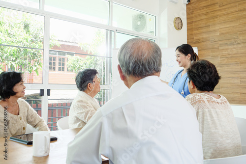 Fototapet Asian healthcare professional in blue scrubs leading discussion with group of senior patients in bright modern clinic, explaining medical care plans and promoting healthy lifestyle awareness