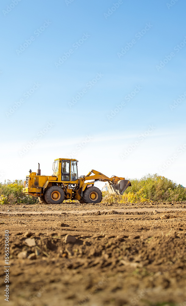 Fototapeta premium Yellow bulldozer at a construction site. Big wheel excavator leveling and clearing the land plot side view. Moving earthworks soil. Copy space. Building area. Special transport. High quality photo.