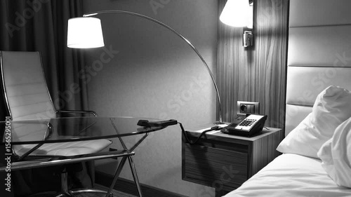 Abstract almost spooky black and white shot of a modern hotel room interior with a old analog phone off the hook on a bedside table, an unmade bed, and a contemporary desk and chair.
