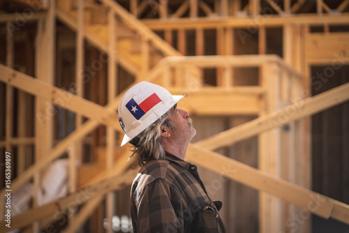 Construction Worker Wearing Hard Hat with Texas Flag Sticker Walking Inside of and Looking Up at Wooden Frame of House at Construction Site