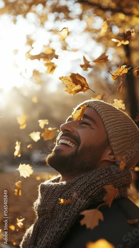 Smiling man enjoying falling autumn leaves wearing beanie and scarf in a park