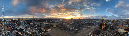 A panoramic view of a vast scrapyard filled with piles of metal and industrial waste under a partly cloudy sky during sunset.