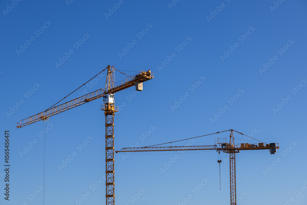 Two large yellow tower cranes stand tall against a clear blue sky, symbolizing active construction, urban development, and industrial infrastructure growth Two large yellow tower cranes stand tall against a clear blue sky, symbolizing active construction, urban development, and industrial infrastructure growth
