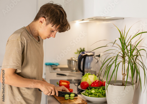 Cheerful satisfied teenage boy preparing by cutting vegetable salad in kitchen