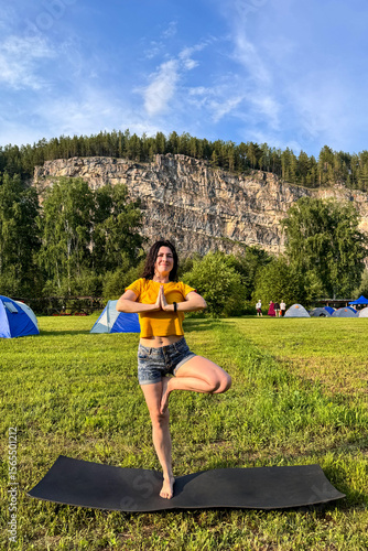 Young woman practices tree pose on a yoga mat in a summer campsite surrounded by blue tents, forest, mountains and open sky, a girl in a tree pose does yoga in nature.