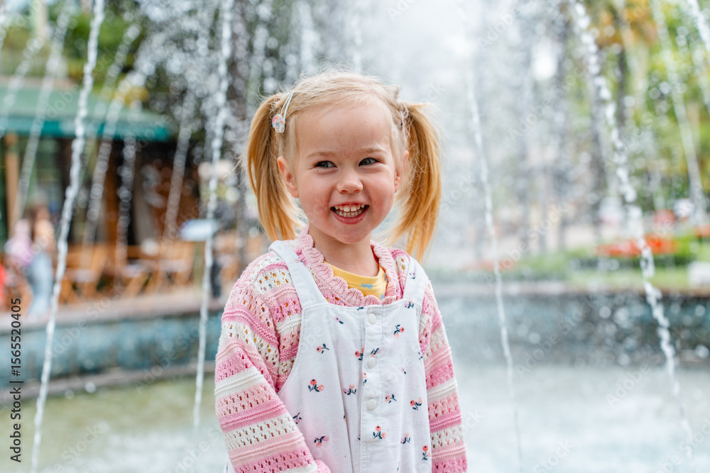 Naklejka premium Child girl laughs and rejoices next to the fountain in the summer garden